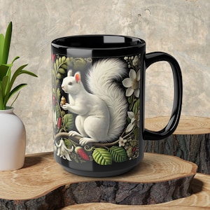 May include: Black ceramic mug with a white squirrel holding an acorn surrounded by green leaves and red berries. The squirrel is on a branch with white flowers.