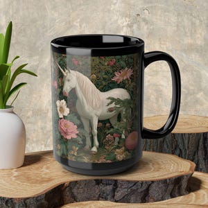 May include: Black ceramic mug featuring a white unicorn in a floral garden scene. The unicorn has a long, flowing mane and a spiraled horn. Pink and white flowers surround the unicorn on the mug's surface. The mug has a large handle.