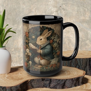 May include: Black ceramic mug featuring a detailed illustration of a rabbit in a blue coat reading a book amidst flowers and foliage. The design has a vintage, storybook aesthetic, with warm colors and intricate details. The mug has a large handle.