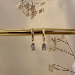 Könnte beinhalten: Ein Paar goldene Creolen mit einem baumelnden rechteckigen, violetten Edelstein.