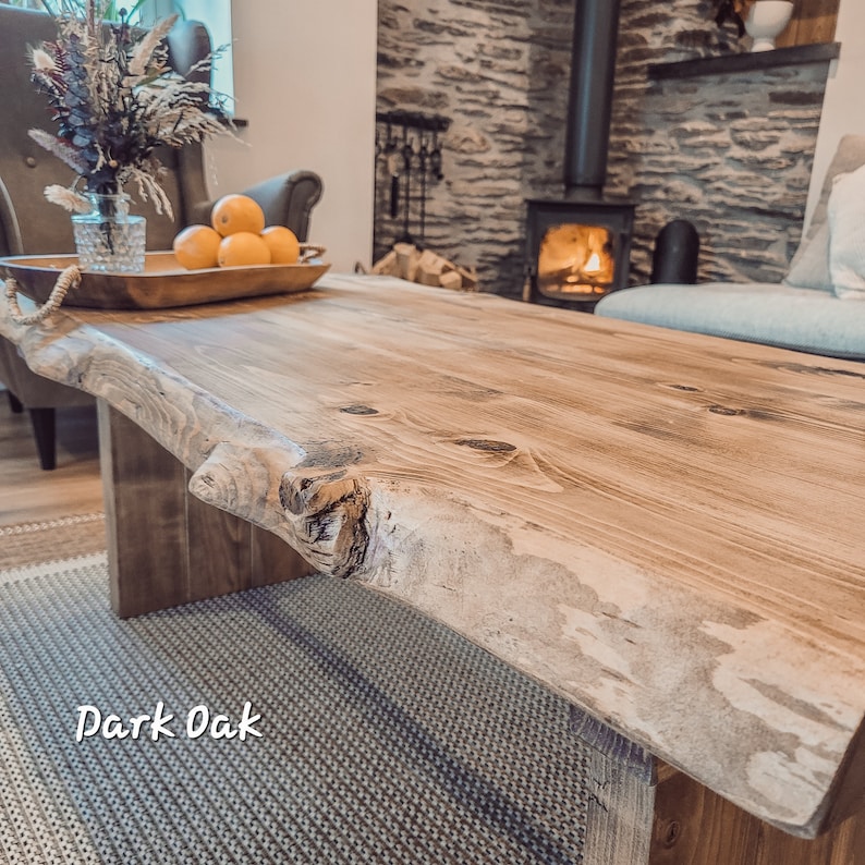 May include: A large, rustic wooden coffee table with a live edge top. The table is made of dark oak wood and has a natural, unfinished look. The table is sitting in a living room with a fireplace in the background.