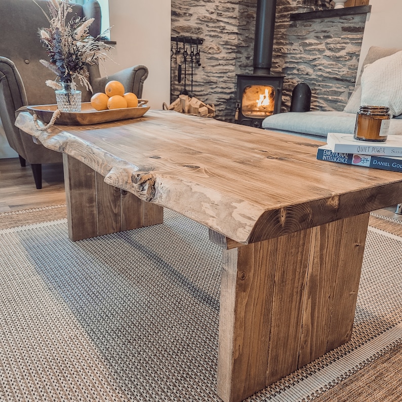 May include: A rustic wooden coffee table with a live edge top and thick legs. The table is made of natural wood with a brown stain and has a rough, unfinished look. The table is sitting on a woven rug in front of a fireplace.