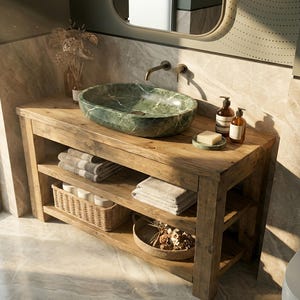 K&ouml;nnte beinhalten: Ein rustikaler Badezimmerschrank aus Holz mit einem ovalen Aufsatzwaschbecken aus gr&uuml;nem Marmor und einem bronzefarbenen Wasserhahn. Das M&ouml;belst&uuml;ck hat zwei offene Regale mit gefalteten grauen Handt&uuml;chern, einem Weidenkorb und einem runden Tablett.