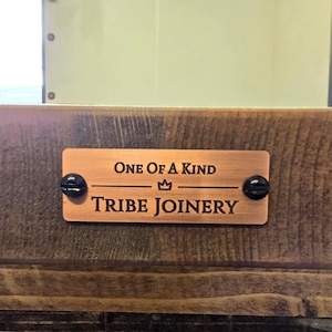 May include: A copper-colored metal sign with black screws reads "ONE OF A KIND" with a crown symbol and "TRIBE JOINERY" in a woodgrain background.