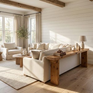 May include: A living room featuring a cream-coloured sofa with a long wooden console table placed behind it, a matching armchair, a round wooden coffee table, and a table lamp with a beige shade, all set against white horizontal shiplap walls.