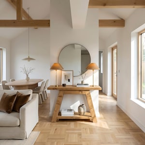 May include: An interior scene featuring a wooden console table with two lamps and a circular mirror. The table displays decorative items. The room includes a sofa, dining table, and parquet flooring. The space is well-lit with natural light.