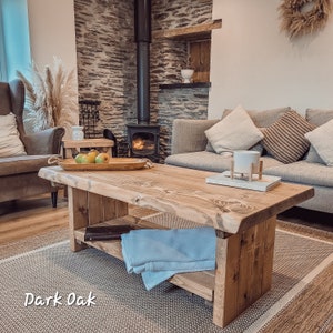 May include: A rustic wooden coffee table with a live edge top and a lower shelf. The table is made of dark oak wood and has a natural finish. The table is sitting on a rug in a living room with a sofa and a fireplace in the background.