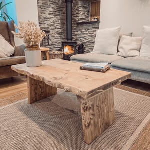 May include: A rustic wooden coffee table with a live edge top and chunky legs. The table is in a living room setting with a couch and a fireplace in the background.
