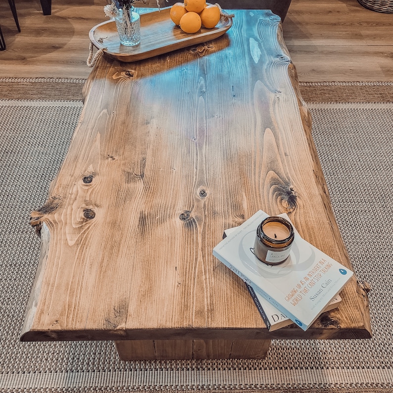 May include: A rustic wooden coffee table with a live edge design. The table has a natural wood finish and is decorated with a bowl of lemons and a book with a candle on top.