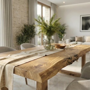 May include: A rectangular dining table featuring a natural live edge wood grain surface with a light beige fabric table runner. A clear glass vase filled with green feathery foliage sits in the centre, accompanied by a small wooden bowl.
