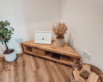 May include: A triangular wooden corner shelf unit with a natural wood grain finish. The unit features an open storage shelf underneath the top surface. A white picture frame, a beige vase with dried grasses, and a woven basket are placed on top.