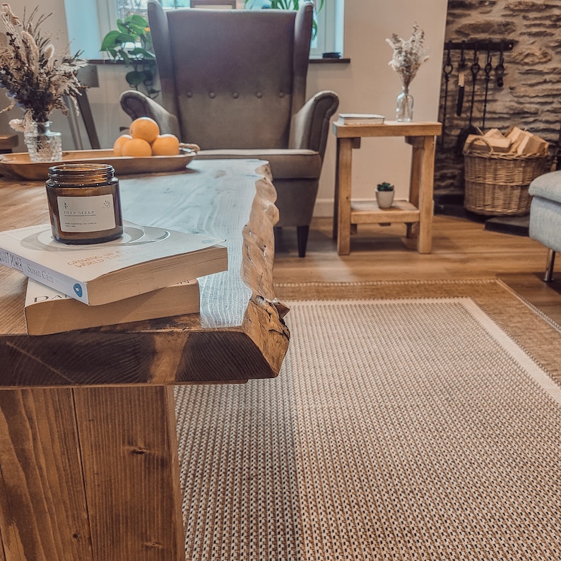 May include: A rustic wooden coffee table with a live edge design. The table has a natural wood finish and is decorated with a candle, a book, and a bowl of oranges. The table is in a living room setting with a rug on the floor.