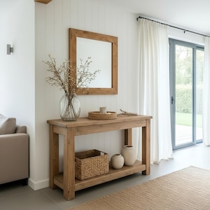 May include: A rustic wooden console table with a framed mirror above. The table features a lower shelf with a wicker basket and two ceramic vases. A glass vase with branches, a woven tray, and decorative items adorn the table's surface.