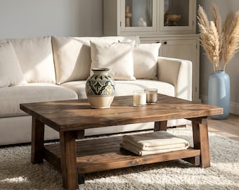 May include: A rustic, square wooden coffee table with a lower shelf, holding a folded blanket. The table is in a living room setting with a cream-colored sofa, decorative vase, candles, and a fluffy rug.