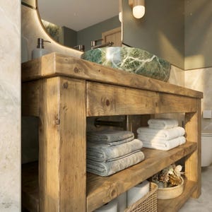 K&ouml;nnte beinhalten: Ein Badezimmerschrank aus hellbraunem Holz mit sichtbarer Maserung und einem gr&uuml;nen Marmor-Aufsatzwaschbecken. Das M&ouml;belst&uuml;ck verf&uuml;gt &uuml;ber ein offenes unteres Regal mit Stapeln gefalteter grauer und wei&szlig;er Handt&uuml;cher sowie einem Korb.