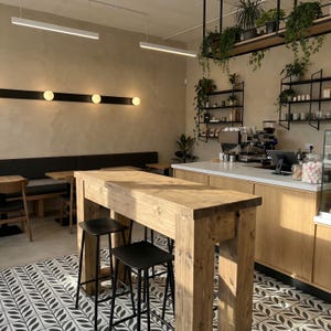 May include: Interior of a coffee shop featuring a large wooden table and black stools. The floor has a black and white leaf pattern. Beige walls, hanging plants, and shelves with coffee-making equipment are also visible.