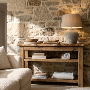 May include: A wooden console table with three shelves. The top shelf has wooden bowls and a lamp with a beige shade. The lower shelves display books, a framed photo, and a folded blanket. The background is a stone wall.
