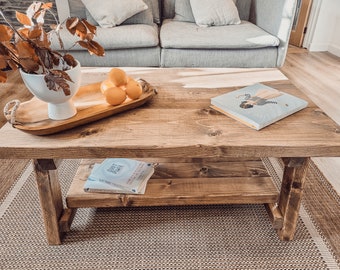 May include: A rustic wooden coffee table with a lower shelf. The table has a natural wood finish and is decorated with a bowl of lemons, a book, and a magazine. The table is sitting on a woven rug.