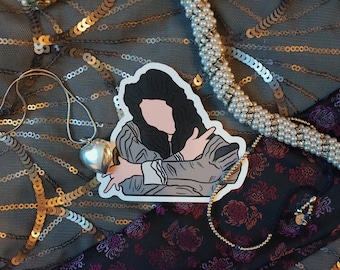 May include: A sticker of a cartoon person with long black hair wearing a grey and white outfit. The sticker is on a background of gold and silver sequins, a black fabric with a floral pattern, and a white pearl necklace.