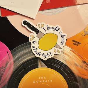 May include: A sticker featuring the phrase "I brought a lemon to a knife fight" and a lemon being cut by a knife. The sticker is placed on a background of vinyl records in various colors.