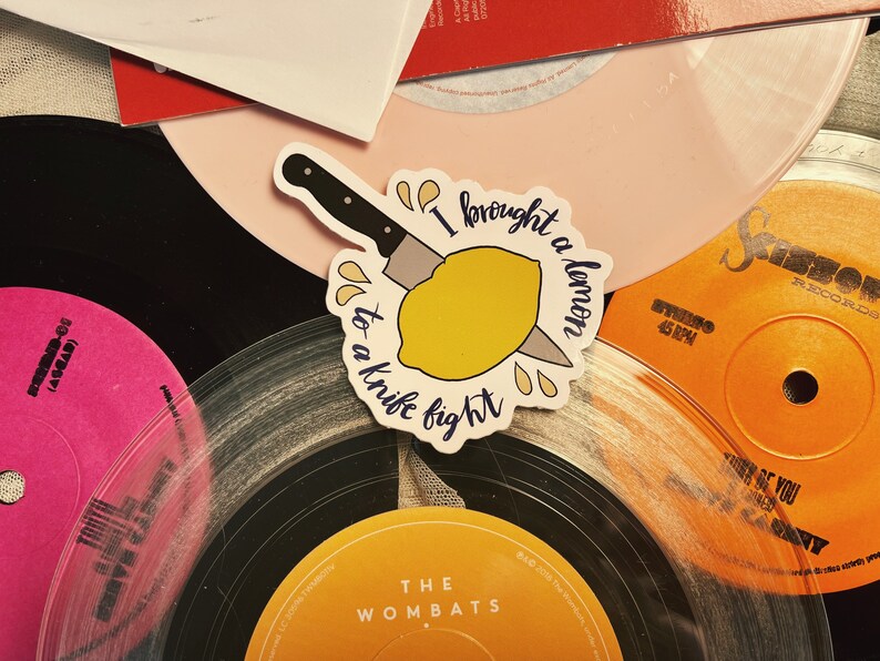 May include: A sticker featuring the phrase "I brought a lemon to a knife fight" and a lemon being cut by a knife. The sticker is placed on a background of vinyl records in various colors.