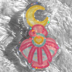 May include: A pink and yellow resin charm with a crescent moon and a heart design. The charm is surrounded by glitter and stars.
