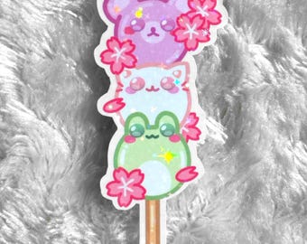 May include: A sticker featuring a stack of three cute animal characters on a stick. The characters are a purple bear, a white cat, and a green frog. Each character is surrounded by pink cherry blossoms.
