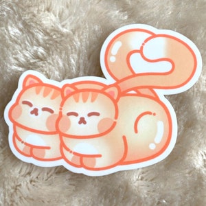 Könnte beinhalten: Ein Aufkleber mit zwei orangefarbenen Katzen, die sich aneinanderkuscheln und ihre Schwänze ineinander verschlungen haben. Die Katzen haben die Augen geschlossen und sind im Cartoon-Stil gezeichnet.
