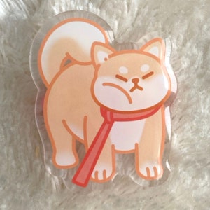Könnte beinhalten: Ein transparenter Acryl-Anhänger mit einem Cartoon-Shiba-Inu-Hund, der einen roten Schal trägt. Der Hund hat einen hellbraunen Körper, weißes Fell an Brust und Schwanz und einen mürrischen Gesichtsausdruck.