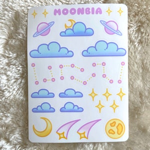Könnte beinhalten: Eine Bogen mit Aufklebern zum Thema Weltraum. Die Aufkleber zeigen Wolken, Sterne, Planeten, Monde und Sternbilder in Pastellrosa, Blau und Gelb. Der Bogen ist mit "MOONBIA" beschriftet.