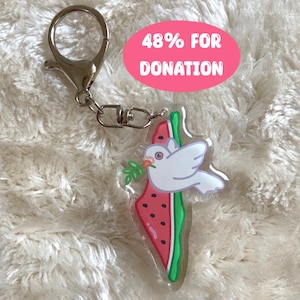 Può includere: Un portachiavi a forma di anguria raffigura una colomba bianca con un ramo di ulivo nel becco. Il portachiavi ha la forma del contorno di Israele. Il portachiavi ha un anello per chiavi argentato. Il testo "48% PER DONAZIONE" è stampato su uno sfondo rosa.