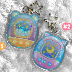May include: Two clear plastic keychains with a star charm. The keychains are shaped like a Tamagotchi, a popular virtual pet toy. One keychain is purple and blue with stars and a crescent moon. The other keychain is orange and yellow with stars and a cloud. The keychains are labeled #1 and #2.