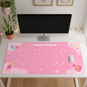 Könnte beinhalten: Eine rosa Schreibtischunterlage mit einem Gittermuster, Sternbildern und dem Text "POWERED BY SPARKLES." Die Unterlage zeigt Cartoon-Illustrationen einer Schleife, eines Buches und anderer himmlischer Elemente. Eine weiße Computermaus liegt auf der Unterlage.
