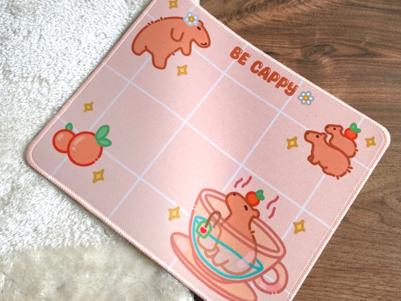 May include: Pink and white mousepad with a tiled design. It features a cartoon capybara in a teacup, two capybaras standing together, and a capybara with a flower on its head. The text "BE CAPPY" is printed on the mousepad.