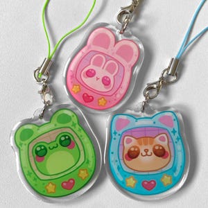 Puede incluir: Tres colgantes de acrílico transparente con diseños de animales de dibujos animados. Uno es un conejo rosa, otro una rana verde y otro un gato azul. Cada colgante tiene un cierre plateado y un cordón de color.