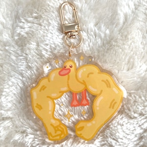 May include: A clear acrylic keyring featuring a yellow duck with orange feet and a pink beak, framed by two cartoon muscular arms. The keyring has a gold-coloured clasp and is decorated with sparkling star accents.