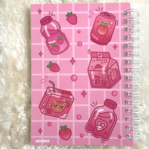 May include: Pink spiral bound notebook with a pink grid pattern and illustrations of pink juice boxes, a pink can, and peaches. The text "MOONBIA" is at the bottom of the notebook.