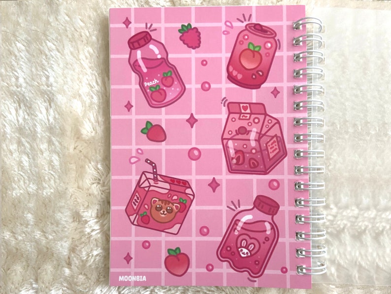 May include: Pink spiral bound notebook with a pink grid pattern and illustrations of pink juice boxes, a pink can, and peaches. The text "MOONBIA" is at the bottom of the notebook.