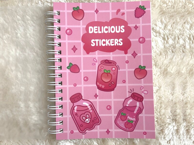 May include: A pink spiral bound notebook with a grid pattern and illustrations of peach-flavored drinks and fruit. The text "DELICIOUS STICKERS" is printed on the cover.