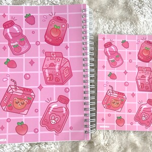 May include: Two pink spiral notebooks with a grid pattern and cute cartoon illustrations of juice boxes, bottles, and fruit. The notebooks have the word "MOONBIA" printed on the bottom left corner.