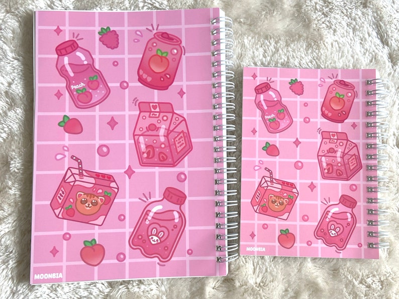 May include: Two pink spiral notebooks with a grid pattern and cute cartoon illustrations of juice boxes, bottles, and fruit. The notebooks have the word "MOONBIA" printed on the bottom left corner.