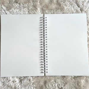 May include: A white spiral bound notebook with a silver binding. The notebook is lying flat on a white fuzzy surface.