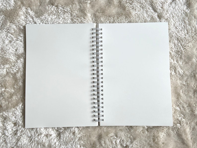 May include: A white spiral bound notebook with a silver binding. The notebook is lying flat on a white fuzzy surface.
