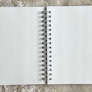 May include: A white spiral bound notebook with a blank page open. The notebook is lying flat on a fuzzy white surface.