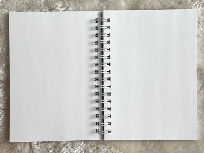 May include: A white spiral bound notebook with a blank page open. The notebook is lying flat on a fuzzy white surface.