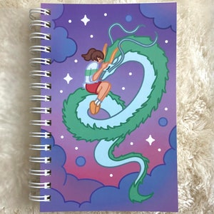 May include: A purple and blue spiral notebook featuring a cartoon illustration of a girl riding a green and blue dragon in a starry sky.