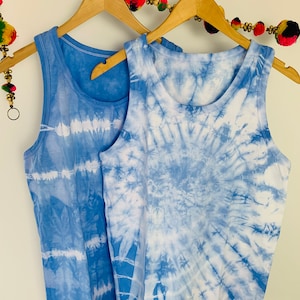 May include: Two sleeveless tank tops hanging on wooden hangers. One tank top features a horizontal striped tie-dye pattern in shades of blue and white, while the other displays a circular spiral tie-dye design in the same blue and white colour palette.