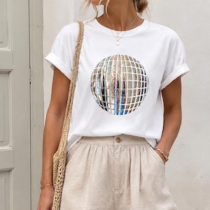 May include: A white short-sleeved t-shirt featuring a central graphic of a silver disco ball with a grid pattern. The shirt is paired with beige high-waisted shorts that have a button closure and a woven straw-style shoulder bag.