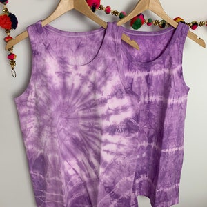 May include: Two purple tie-dye tank tops hanging on wooden hangers. The tank tops have a white background with purple swirls and stripes.