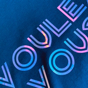 May include: Navy blue t-shirt with the word "VOUS" in a holographic, iridescent design. The text is angled across the shirt, creating a modern and eye-catching look. The shirt is made of a soft fabric.